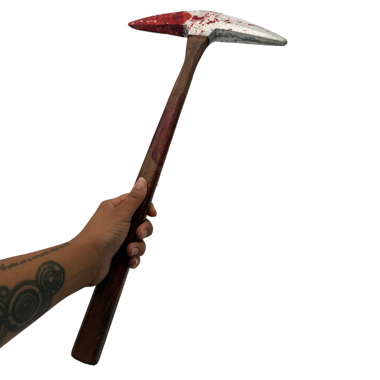 A hand holding a toy or prop pickhead hammer with a dark brown wooden handle and a metal head splattered with red paint, isolated on a white background.