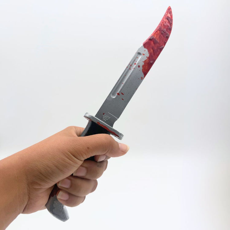 In hand view 12 inch plastic survival hunting knife inspired by the 120 Buck design from the Scream horror movie franchise, featuring a silver blade and black handle, blood on knife tip and handle, designed for cosplay or display