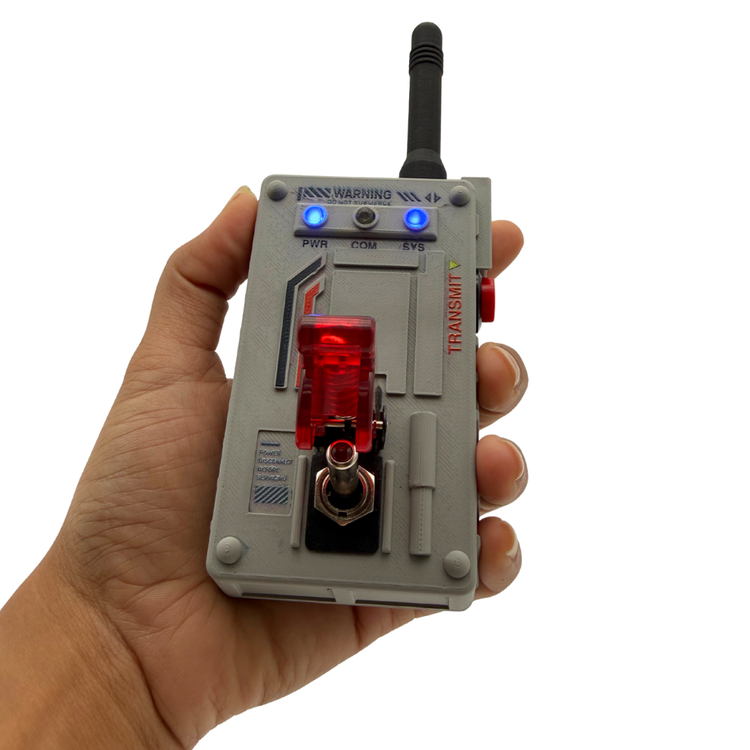 A hand holding a gray electronic transmitter device with a black antenna, blue indicator lights, a red translucent switch, and printed technical labels on a white background.